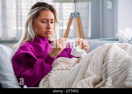 Maladie, virus saisonnier concept de problème. Femme malade ayant la grippe allongé sur un canapé regardant la température sur un thermomètre. Femme malade couché au lit avec Banque D'Images