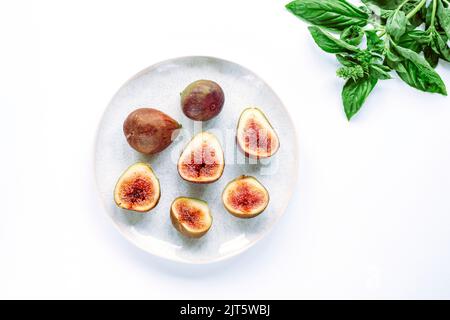 Figues mûres fraîches sur une assiette et feuilles de basilic sur une table blanche. Vue de dessus, plan d'appartement. Banque D'Images