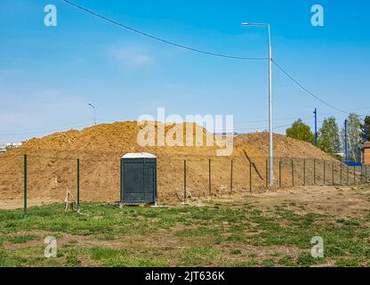 Travaux de terrassement en extérieur avec une grande pile de sable derrière la clôture et une cabine de toilettes extérieures. Banque D'Images