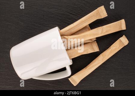 Plusieurs bâtonnets de sucre dans des sacs en papier avec une tasse blanche sur une pierre d'ardoise, vue rapprochée, vue du dessus. Banque D'Images