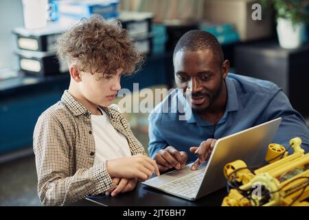 Portrait d'un enseignant noir aidant un jeune garçon à construire un robot pendant les cours d'ingénierie à l'école et utilisant un ordinateur portable pour la programmation ensemble Banque D'Images