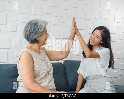 Jeune femme asiatique, fille joyeuse donnant cinq hauts à sa mère, vieille femme, assis sur un canapé gris dans la chambre blanche. Mère et fille cele Banque D'Images