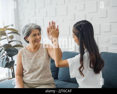 Femme asiatique âgée, maman donnant cinq hauts à sa fille gaie, jeune femme de beauté, assis sur un canapé gris dans la pièce blanche. Mère et fille Banque D'Images