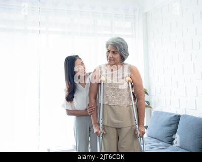 Femme âgée essayant de marcher sur des béquilles debout et maintenues dans les bras par de jeunes femmes asiatiques soigneusement dans la salle de récupération, aidant les vieilles femmes, guérir Banque D'Images