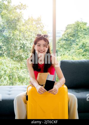 Jeune femme asiatique très heureuse avec lunettes de soleil tenant passeport et billet d'avion avec sourire, asseyez-vous sur le canapé près de chapeau de plage, valise jaune verticale s. Banque D'Images