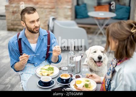 Un homme et une femme élégants ont un petit déjeuner tout en étant assis avec un chien dans un café à l'extérieur Banque D'Images