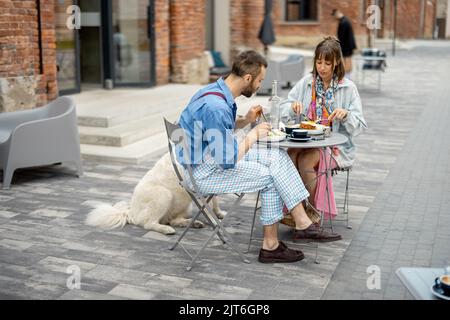 Un homme et une femme élégants ont un petit déjeuner tout en étant assis avec un chien dans un café à l'extérieur Banque D'Images