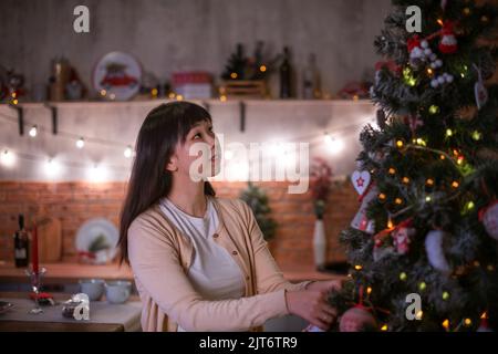 Une jolie fille coréenne aux cheveux sombres dans une confortable salle de Noël avec des lumières de bokeh festives en soirée habille un sapin de Noël avec des jouets écologiques. Banque D'Images