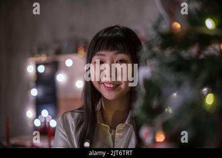 Jolie fille coréenne aux cheveux sombres dans une confortable salle de Noël avec des lumières de bokeh festives le soir. Portrait. Banque D'Images