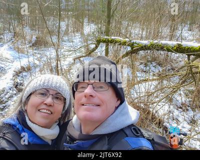 Couple prenant un selfie contre la végétation sans feuilles et la neige au sol, regardant vers le haut, sourire, grand sourire, lunettes, vêtements d'hiver, serre-tête en crochet, bla Banque D'Images