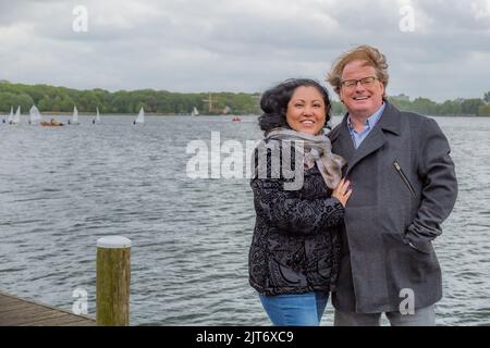 Portrait d'un couple adulte souriant et heureux d'âge moyen contre les eaux d'un lac avec des bateaux à voile naviguant dans le fond flou, hiver nuageux Banque D'Images