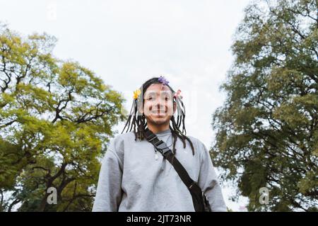 latin vénézuélien jeune homme avec des dreadlocks et des fleurs sur ses cheveux debout dans le parc timide et souriant regardant la caméra, avec des arbres et le ciel Banque D'Images