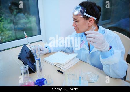 Une femme expérimentée multiethnique, pharmacienne scientifique en blouse de laboratoire blanche, écrit des notes de ses recherches en pharmacologie Banque D'Images