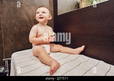 Portrait d'un bébé heureux avec un peigne dans la salle de bains près du miroir. Bébé garçon assis sur une serviette. Enfant d'un an Banque D'Images