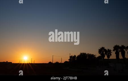 Coucher de soleil en Sicile. Le ciel passe du bleu à l'orange. Les rayons du soleil brillent. Les paumes et autres arbres fournissent de l'ombre entre les câbles d'alimentation et les pylônes. Banque D'Images