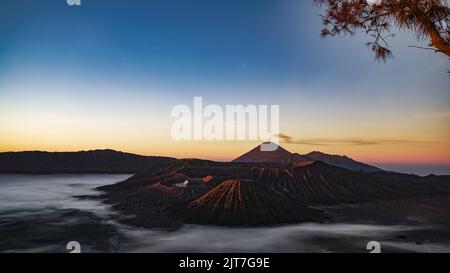 Vue panoramique mystique et surréaliste sur les montagnes des cratères du volcan Bromo au lever du soleil avec ciel bleu et orange clair et reflet de la lumière du soleil et brumeux g Banque D'Images