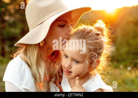 Portrait de famille aimante dans la forêt illuminée par le coucher du soleil en été. Jeune femme mère embrassant temple de petite fille. Banque D'Images