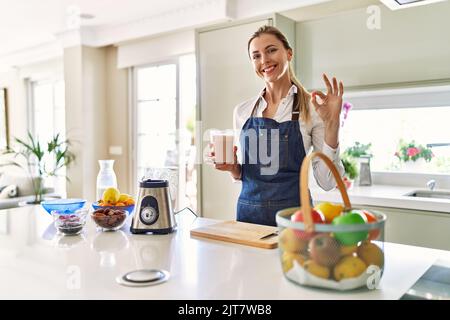 Belle femme blonde portant un tablier buvant smoothie à la cuisine faisant signe ok avec les doigts, souriant sympathique gestante excellent symbole Banque D'Images