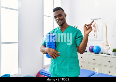 Un jeune physiothérapeute africain tenant un rouleau en mousse à la clinique souriant se réjouit de pointer avec la main et le doigt sur le côté Banque D'Images