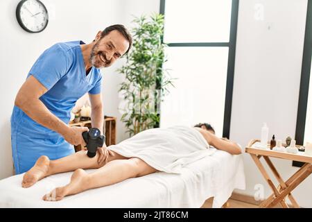 Homme et femme d'âge moyen portant un uniforme de thérapeute ayant une séance de massage des jambes à l'aide d'un pistolet à percussion au centre de beauté Banque D'Images