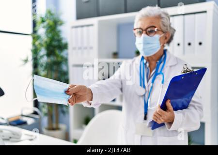 Femme sénior aux cheveux gris portant l'uniforme du médecin donnant un masque médical à la clinique Banque D'Images