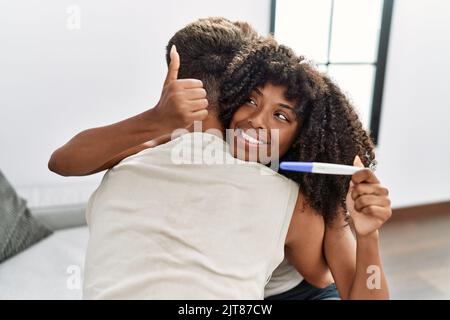 Jeune couple interracial tenant le résultat du test de grossesse pointant le pouce vers le côté souriant heureux avec la bouche ouverte Banque D'Images