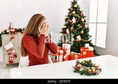 Jeune femme blonde caucasienne assise sur la table par arbre de noël avec une expression triste couvrant le visage avec les mains en pleurant. Concept de dépression. Banque D'Images