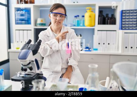 Fille hispanique avec le syndrome de Down travaillant au laboratoire scientifique pointant avec le doigt de la main sur le côté montrant la publicité, sérieux et visage calme Banque D'Images