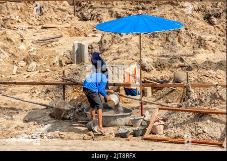 Un ouvrier de la construction verse un seau de petites pierres dans un récipient de mélange de béton tandis que l'autre les mélange. Banque D'Images