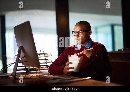 La nourriture est la clé du succès des quarts de nuit. Un beau jeune homme qui mange de la nourriture chinoise tout en travaillant tard dans son bureau. Banque D'Images