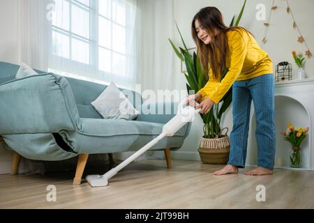 Femme de ménage nettoyage de la poussière sol sous canapé ou mobilier de canapé avec aspirateur, jeune femme asiatique heureuse avec accumulateur aspirateur à hom Banque D'Images