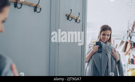 Une cliente féminine attirante choisit un manteau élégant dans la cabine d'essayage, regarde le grand miroir et vérifie le tissu et la qualité. Elle touche le vêtement avec plaisir et sourire. Banque D'Images