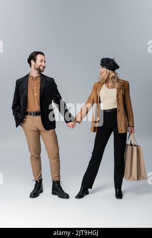 homme heureux tenant les mains de la femme blonde dans un béret élégant debout avec des sacs de shopping sur gris, image de stock Banque D'Images