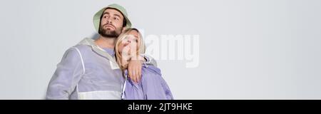 homme barbu au panama chapeau embrassant blonde petite amie en manteau de pluie violet isolé sur gris, bannière, image de stock Banque D'Images