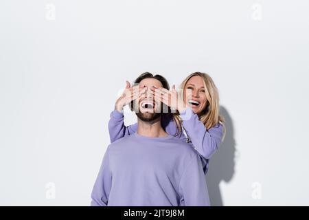 bonne femme blonde couvrant les yeux de l'homme barbu en sweatshirt violet sur gris, image de stock Banque D'Images