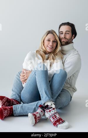pleine longueur de couple gai en tenues d'hiver et chaussettes rouges avec ornement assis sur gris, image de stock Banque D'Images