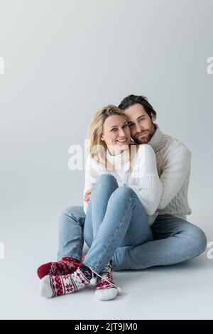 pleine longueur de couple heureux en tenues d'hiver et chaussettes rouges avec ornement assis sur gris, image de stock Banque D'Images