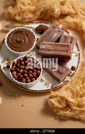 Des pop-glaces au chocolat et aux noisettes servies avec du chocolat fondu sur une assiette blanche. Vue de dessus sur fond beige. Banque D'Images