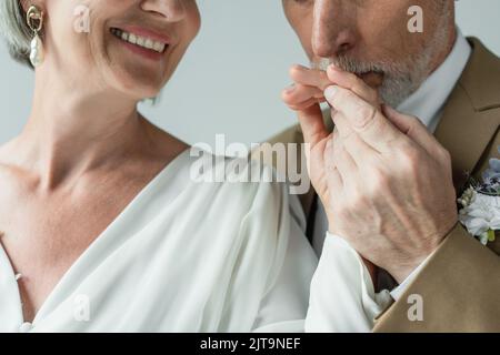 vue rognée de l'homme d'âge moyen barbu embrassant la main de la mariée heureuse en robe de mariage blanche isolée sur gris, image de stock Banque D'Images