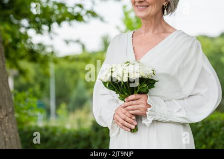 vue rognée de la mariée heureuse d'âge moyen en robe blanche tenant le bouquet de mariage dans le jardin vert, image de stock Banque D'Images