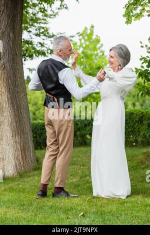 heureux homme mature en tenue formelle dansant avec mariée en robe de mariage blanche dans le jardin vert, image de stock Banque D'Images