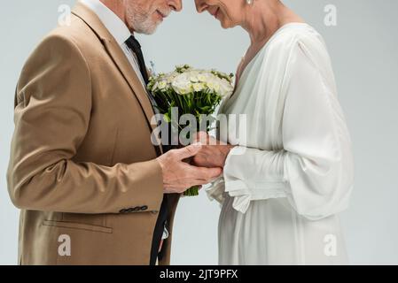 vue rognée du marié d'âge moyen et de la mariée heureuse en robe blanche tenant les mains près du bouquet de mariage sur gris, image de stock Banque D'Images