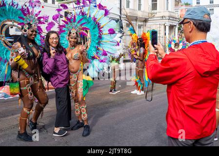 Notting Hill, Londres, Royaume-Uni. 29th août 2022. Le plus grand festival de rue d'Europe est revenu dans les rues de Notting Hill après les années annulées en raison de la pandémie de Covid. Des danseurs exotiques et des groupes musicaux sur le thème jamaïcain défileront dans les rues, avec de la nourriture et des divertissements dans la rue autour de la région, ajoutant à l'événement. Le Grand Parade a lieu le lundi des fêtes de la Banque comme point culminant du festival de trois jours qui a commencé en 1966. Les participants se rassemblent à proximité dans leurs costumes colorés. Visiteurs asiatiques prenant une photo avec des participantes en costumes aux couleurs vives Banque D'Images