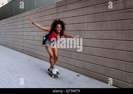 Adolescente multiraciale à cheval sur un skateboard devant un mur en béton, en équilibre. Vue latérale. Banque D'Images