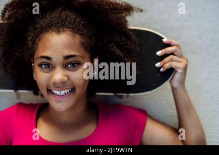 Portrait d'une adolescente multiraciale avec planche à roulettes, devant un mur en béton pendant la journée d'été. Banque D'Images