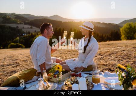 Couple séduisant amoureux, pique-nique et vin à boire sur la colline au coucher du soleil. Banque D'Images