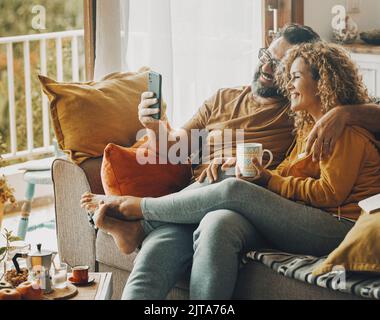 Charmant couple caucasien souriant assis sur un canapé à la maison prenant le petit déjeuner ensemble tout en utilisant le téléphone portable Banque D'Images