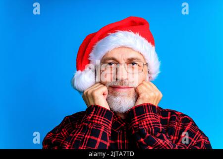 un homme âgé et bien entretenu dans une chemise à carreaux et un chapeau de père noël dans un fond de studio bleu Banque D'Images