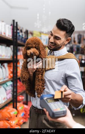 Homme d'arabie souriant qui regarde la caniche tout en payant avec une carte de crédit dans la boutique pour animaux Banque D'Images