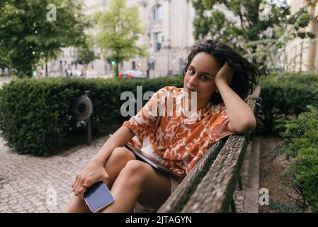 jeune femme en tenue d'été tenant smartphone et assis sur un banc dans parc vert, image de stock Banque D'Images